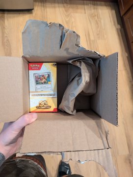 A person's tattooed hand holds open a cardboard shipping box containing crumpled gray packing paper and a Pokémon trading card game booster box.