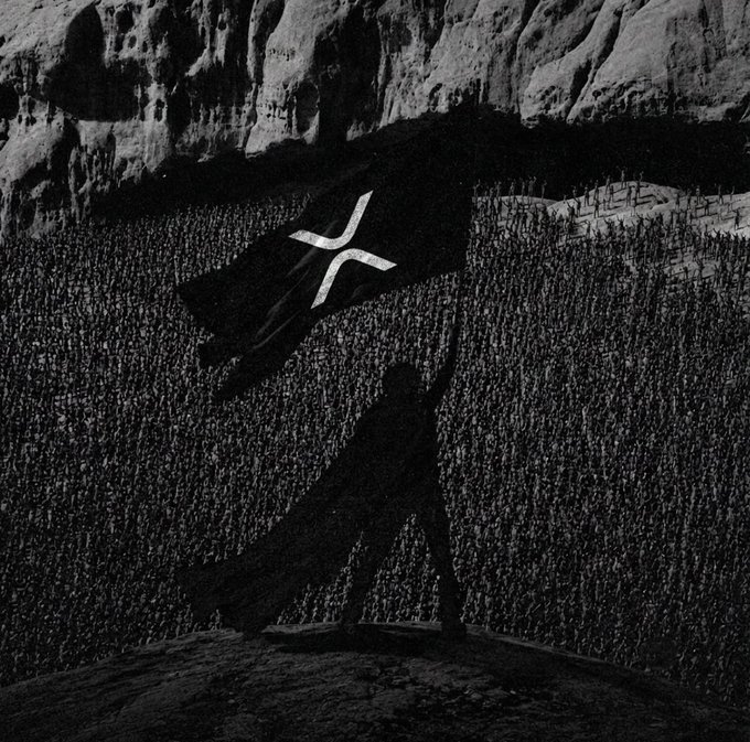 The image features a person holding up a flag with an X on it, which appears to be a symbol of cryptocurrency called Ripple. The flag is being raised above their head while they stand on top of a rock or cliff. This suggests that the person might be promoting Ripple as a means of payment or supporting its use in the digital currency market.