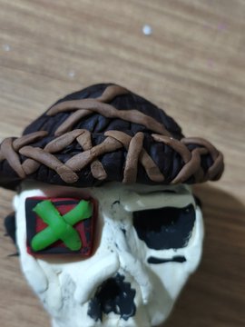 A handmade clay skull wearing a brown woven hat rests on a wooden surface, featuring a green "X" symbol painted on its forehead. The post highlights the creator's dedication to finishing the piece late at night for an art showing, emphasizing that consistency is key to gaining support.