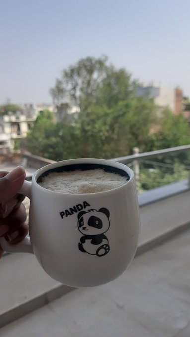 A hand holds up a white mug with a panda graphic and the word 'PANDA', filled with a frothy beverage, against a blurred background of trees and a balcony.