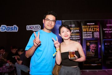 A young couple poses with peace signs at a crypto-themed event, with banners in the background advertising 'Dash', 'LIQUID', and 'BUILD CRYPTO WEALTH' and 'CRYPTO REGRET'.