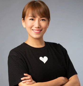 A smiling Asian woman with short brown hair wearing a black t-shirt with a white heart design, posing with arms crossed against a gray background.