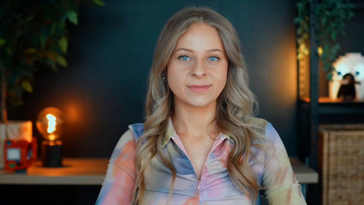 The image features a woman with blonde hair wearing a colorful shirt, sitting at a desk or table. The background includes a dark wall and a lamp that casts light onto her face as she looks directly into the camera. There are also some books on the table in front of her.