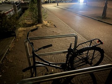 A nighttime photo of a black bicycle locked to a metal railing on a sidewalk next to a street.