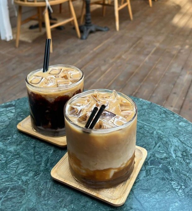The image shows two glasses of coffee sitting on a table, each accompanied by a straw. The cups are placed on wooden coasters, adding an elegant touch to the scene. In the background, there are chairs and dining tables, suggesting that this is likely taking place in a restaurant or cafe setting. A caption for the image reads: "Good afternoon CT @useTria provides a single interface for all chains. You can accumulate XP and enjoy cash back directly with Tria cards."