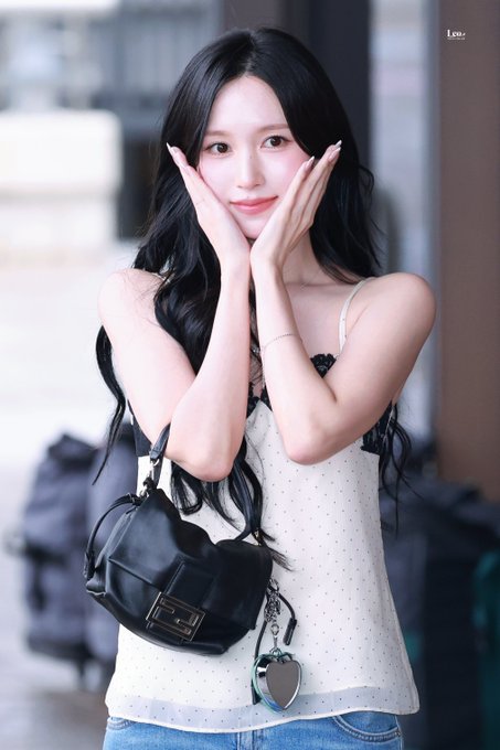 The image features a young woman with long black hair posing with her hands on her cheeks, wearing a white polka-dot top and carrying a black heart-shaped bag. A small "tw" watermark appears in the top right corner, identifying her as Mina from the K-pop group TWICE, consistent with the hashtags in the accompanying post.