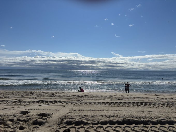 A serene beach scene with two people near the shoreline, sandy beach in the foreground showing tire tracks, ocean waves, and a partly cloudy blue sky with sunlight reflecting on the water.