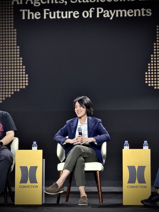 A woman in a navy blazer and olive trousers sits holding a microphone on a panel stage beneath a screen reading 'AI Agents, Stablecoins & The Future of Payments,' flanked by yellow 'CONVICTIION' branded podiums.