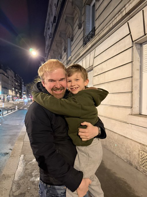 A man with reddish-blonde hair and beard, wearing a black puffer jacket, kneels on a nighttime city street to share a warm, smiling embrace with a young boy in an olive green hoodie. The tender father-son moment, set against a Parisian-style building backdrop, reflects the post's theme of belonging and pride in being part of a close-knit, meaningful community.
