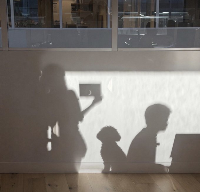 Shadows cast on a beige wall and wooden floor showing two people and a small dog; one silhouette appears to be holding up a phone as if taking a picture.