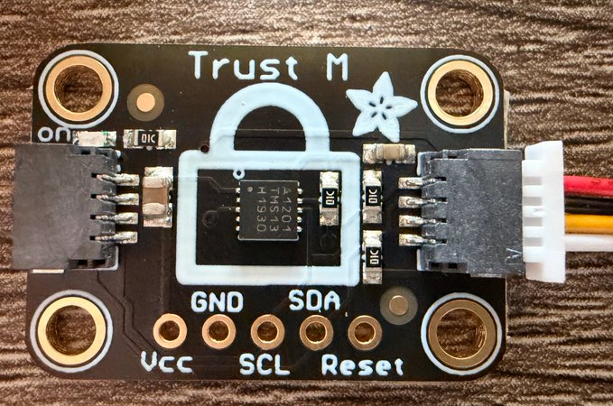 A black circuit board labeled "Trust M" featuring a lock icon, star design, and a visible chip marked "ATSHA204A H133T" with exposed pins for GND, SDA, VCC, SCL, and Reset connections, along with red, yellow, and black wires attached. The image shows the hardware wallet mentioned in the post—a small, affordable security chip module that the author suggests players will need to obtain in order to participate in an upcoming game requiring verifiable hardware wallets.