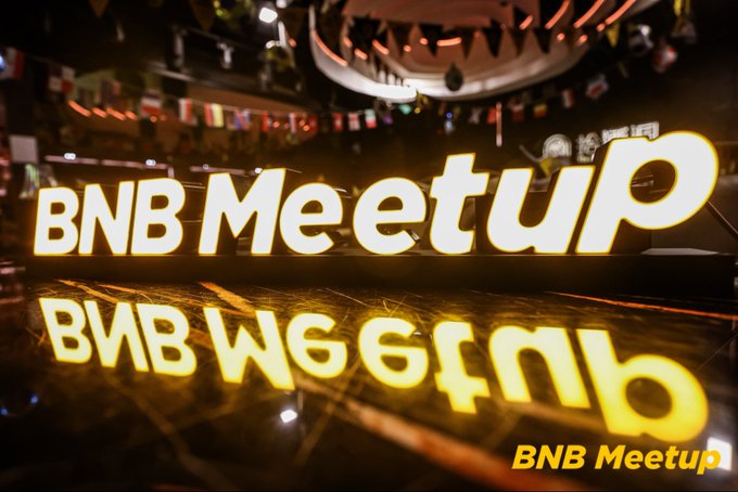 A photograph of an illuminated 'BNB Meetup' sign with mirrored Georgian script below, set in a venue with international flags hanging from the ceiling.