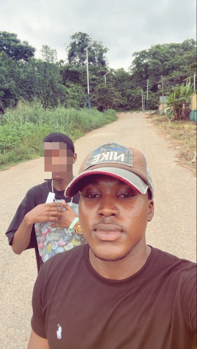 A selfie of two young men standing on a dirt road surrounded by lush green vegetation, with the person in front wearing a Nike cap and brown shirt.