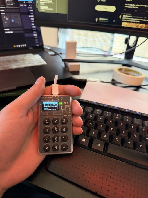 A hand holds a Trezor "Colossus" hardware wallet displaying "Don't trust me, verify yourself" on its screen, set up next to a keyboard and a laptop featuring the "teslaonthebeach" website. The image illustrates the post's message about taking the contrarian step of finally setting up self-custody crypto storage rather than relying on centralized exchanges.