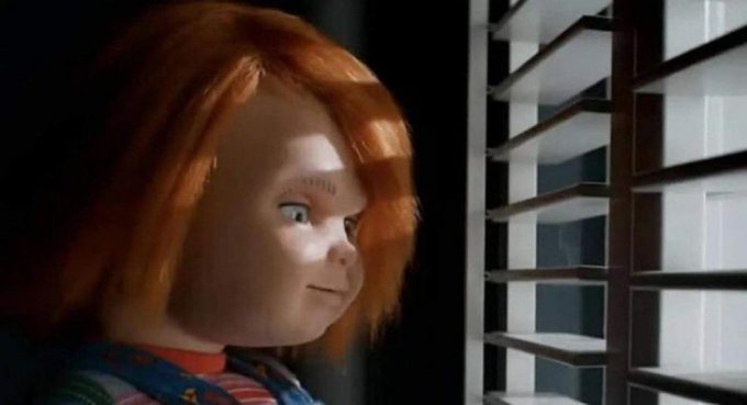 A close-up of a Chucky doll from the horror franchise, showing its red hair and stitched face, peeking through white window blinds.