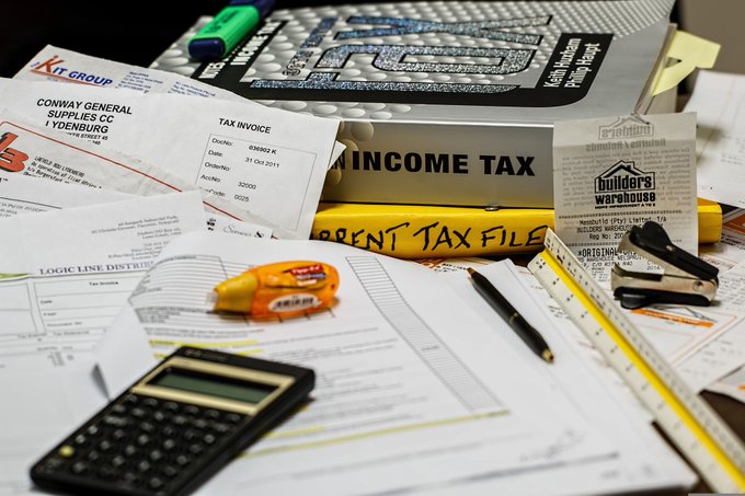 A cluttered desk scene featuring tax-related documents, including a 'TAX INVOICE' from 'CONWAY GENERAL SUPPLIES CC', an 'INCOME TAX' file folder, a calculator, pens, a ruler, and a tape measure.