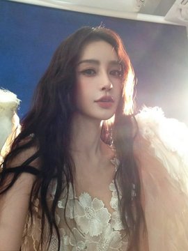 A woman with long dark hair and delicate makeup wearing a white lace top with feathery wings, photographed in soft lighting.