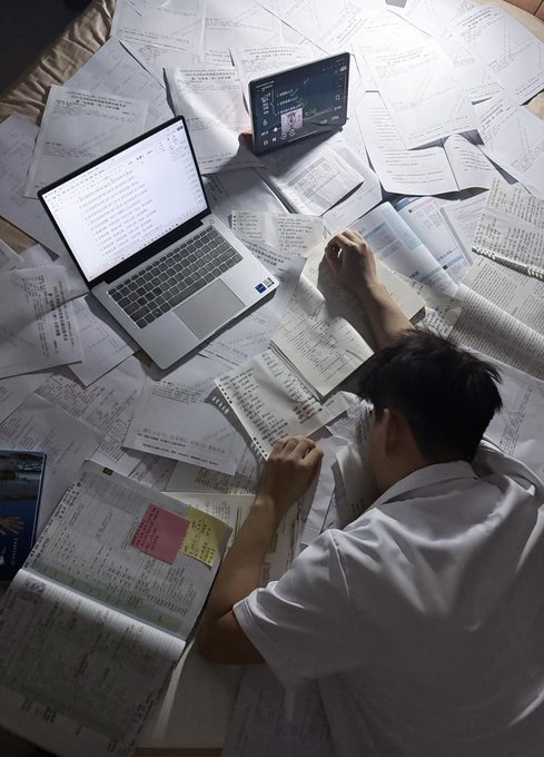 A student lies on a bed buried under countless printed papers and handwritten notes, typing on a laptop with another screen propped up nearby, visually capturing the overwhelming burden of manually gathering and processing real-world data—a stark contrast to the AI post's point that embodied AI systems and world models fundamentally depend on vast amounts of such grounded, physical-world data to function.