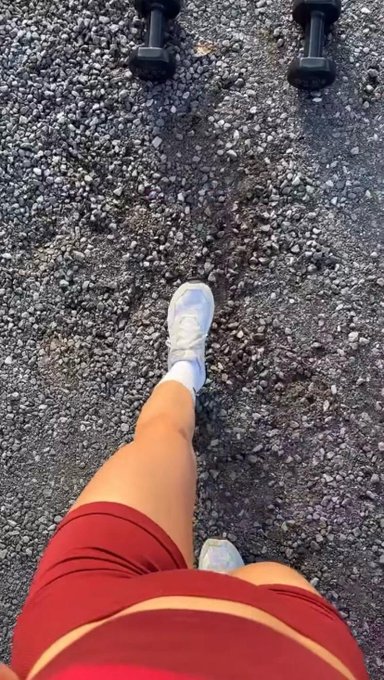 A first-person view looking down at legs in maroon shorts and white sneakers walking on gravel, with dumbbells visible at the top of the frame.