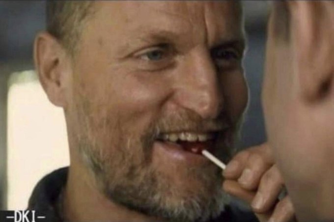A close-up scene from the film 'Druk' (seen by the '—DK1—' watermark) showing a bearded man smiling while holding a small white object near his mouth, facing another person.