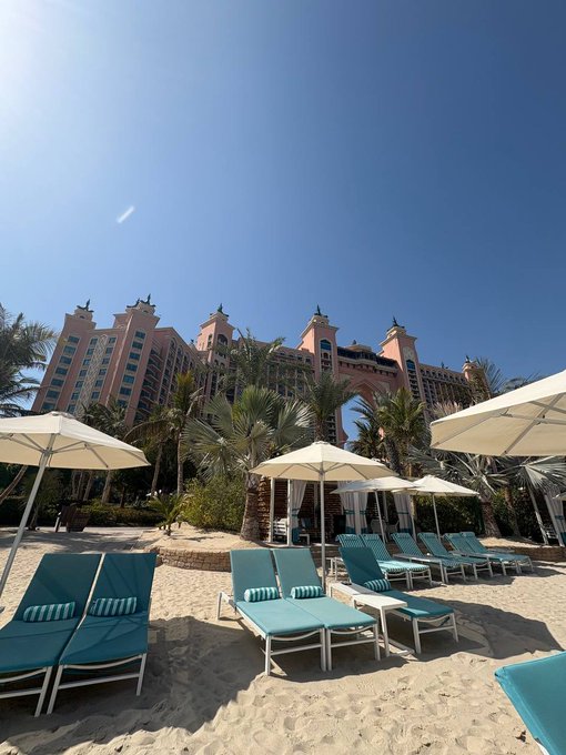 A beach resort scene with turquoise lounge chairs and white umbrellas on sandy ground, with the distinctive pink Atlantis hotel building and palm trees in the background under a clear blue sky.