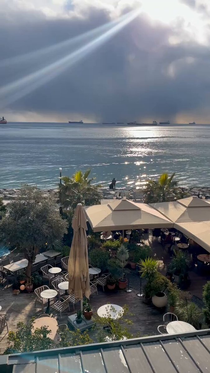 The image shows a beautiful beach scene with an ocean view, palm trees, umbrellas, tables, chairs, and potted plants. The sun is shining brightly through the clouds, creating a warm and inviting atmosphere. In the background, there are several boats visible on the water. The post features a photo of a person's face that says "gm legends make sure to touch grass and check out @sleepagotchi". This adds an interesting detail to the image, making it more engaging for viewers who might be interested in the person's story or the location they are sharing their experience at.