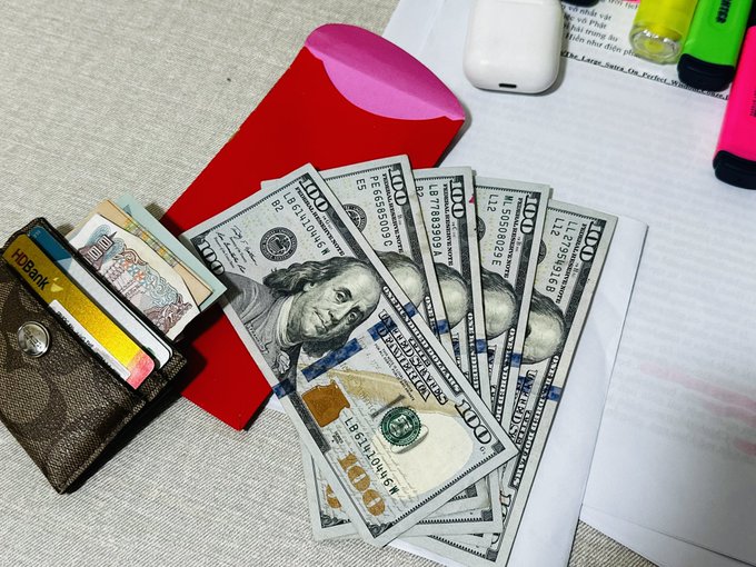 A fanned-out spread of US $100 bills on a desk next to a brown wallet, a red envelope, an AirPods case, and highlighters.