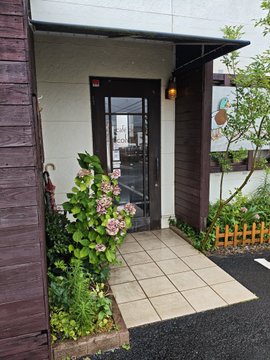 A small renovated shop entrance with a paved walkway, lush hydrangeas, and a hanging ornament under an awning reflects the post's message of diligently repairing old storefronts, inviting JPYC customers to visit despite the rainy morning.