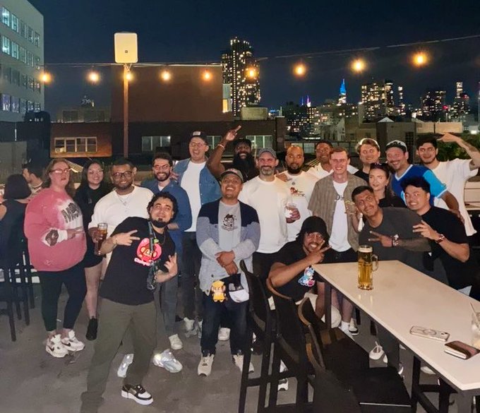 A group of people pose for a photo at a rooftop gathering at night with string lights overhead and a city skyline visible in the background.