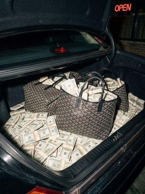 The open trunk of a car is filled with stacks of bundled US dollar bills, with two designer handbags resting on top of the money.