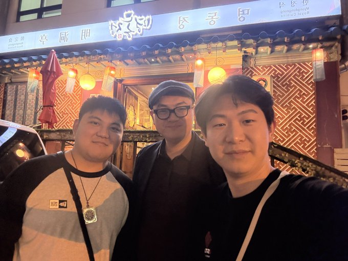Three men pose for a photo outside a Korean restaurant at night with signs in Korean and Chinese characters, including '두공방' and '喜 丽門', with an American Eel House sign visible.