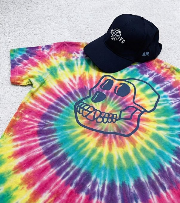 A vibrant rainbow tie-dye t-shirt featuring a navy blue Bored Ape Yacht Club (BAYC) skull graphic is displayed on a white surface, paired with a black dad cap branded with a globe logo and "VC" lettering, matching the post's showcase of bold BAYC merchandise.