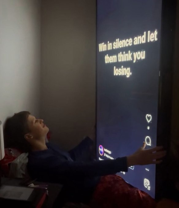 A young boy reclines in a dark room while looking up at a large screen displaying the motivational quote "Win in silence and let them think you losing."