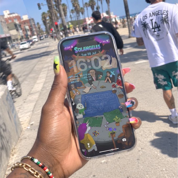 A hand with neon green nails displays a smartphone showing a custom "SOLANGELES" lock screen with the time 16:02 and crypto tickers (BTC, ETH, SOL) on a sunny Los Angeles boardwalk, teasing the upcoming Solangeles NFT series drop.