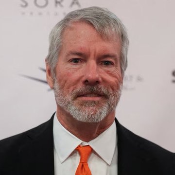 The image features a man wearing an orange tie with white collar and black suit jacket. The background of the photo shows a white wall with a logo that reads "SARA". There are no other objects or people visible in the picture, and the focus is on the man's attire and the surrounding environment.