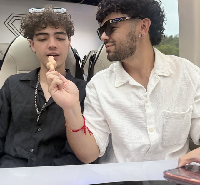 The image shows two men sitting at a table, with one man holding an ice cream cone while the other man looks on. The background features a car and a cell phone, suggesting that they might be enjoying their treat outdoors or during a road trip. The caption "focus on what is in the image and how it relates to this post" implies that the image could be used as part of a social media post promoting a product or service related to cars or road trips.