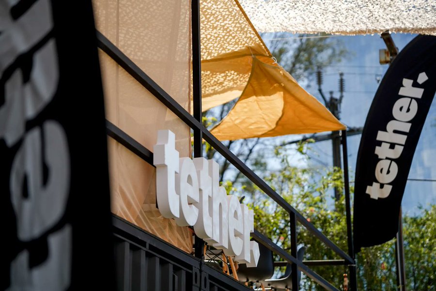 The "tether" logo and branded flag are displayed prominently outside a venue with a beige canopy, tying directly to the Senate investigation highlighting Tether's USDT stablecoin as a key tool in Iran's sanctions-evasion network. The corporate branding in the image underscores the central role of Tether in the alleged financial scheme described in the post.