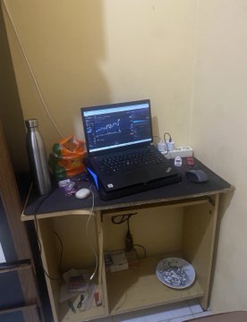 A laptop displaying trading charts set up on a simple wooden desk alongside a water bottle, with power strips and cables visible on the lower shelf.