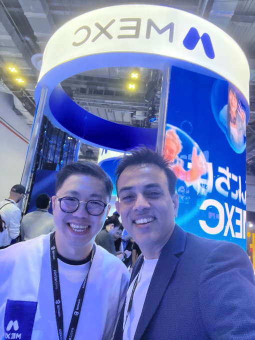 Two smiling men pose for a selfie at what appears to be a trade show, with an MXC exchange booth visible in the background.