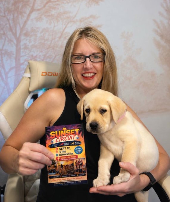 A smiling woman with glasses holding a Labrador puppy and a flyer for 'Sunset Circuit' event on Sept 12 at 5 PM at Oceanfront Park.