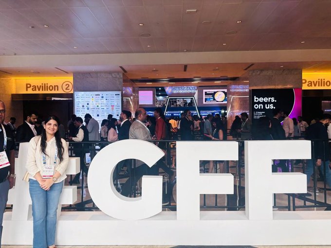 A young woman standing next to large white '#IGFF' letters at the IGFF (International Government Financial Forum) event, with 'Pavilion 2' signage and digital screens visible in the background.