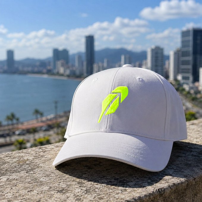 A white baseball cap featuring a bright neon green feather logo sits on a stone ledge, overlooking a coastal city skyline with a beach and high-rise buildings in the background. The feather emblem mirrors the iconic Robinhood brand identity, tying into the post's shoutout to those building with @RobinhoodCrypto.