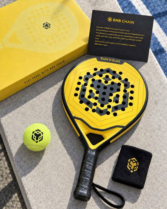 A BNB Chain padel gift set is displayed on a stone surface, featuring a yellow and black paddle branded "Build N Build" alongside a yellow tennis ball, wristband, and a yellow box labeled "PLAY PADEL WITH BNB CHAIN." A thank-you card from the BNB Chain Team accompanies the set, celebrating six years of the blockchain and expressing gratitude to the community. This aligns with the post highlighting branded padel merchandise gifted to BNB Chain community members.