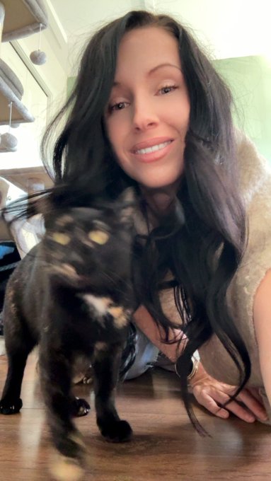 A woman with long dark hair smiles for a selfie alongside a small black dog in what appears to be a kitchen setting.
