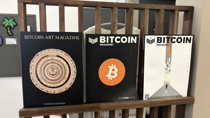 Three magazines displayed on a wooden rack: "BITCOIN ART MAGAZINE GENESIS EDITION" with a circular metallic design, "BITCOIN MAGAZINE THE CORE ISSUE" with an orange logo, and another "BITCOIN MAGAZINE" issue featuring geometric artwork.