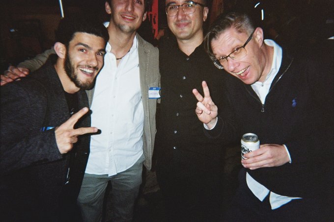 Four men posing together at what appears to be an event, with one holding a beer can and others making peace signs.