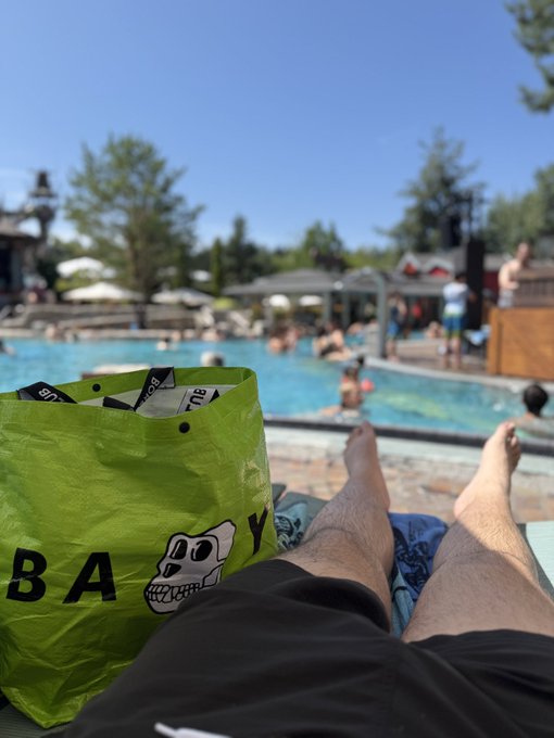 A first-person view of legs relaxing by a crowded outdoor pool on a sunny day, with a bright green tote bag featuring a dog graphic in the foreground.