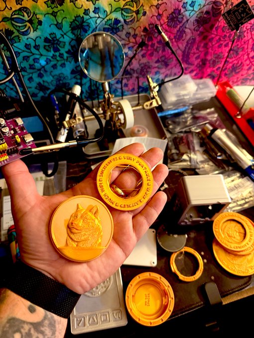 A person's hand holding several gold coins, including one with a Shiba Inu dog design and another with a ring and runes around the edge, with crafting equipment visible on the workbench behind.