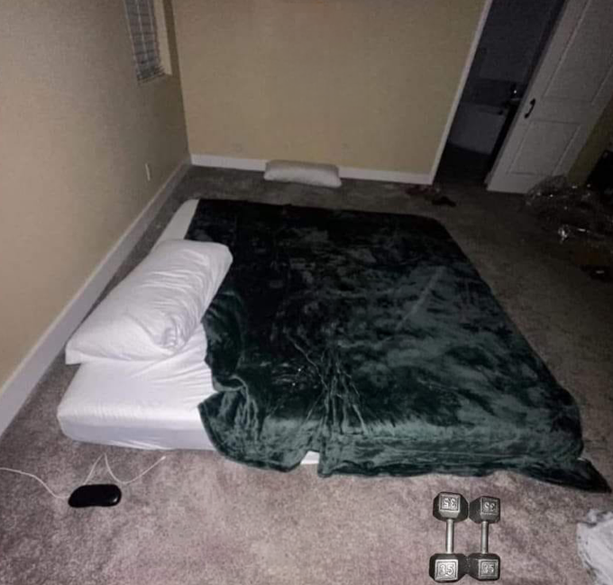 A bedroom floor setup with a mattress, white pillow, and dark green blanket, with two 25-pound dumbbells and a phone nearby.