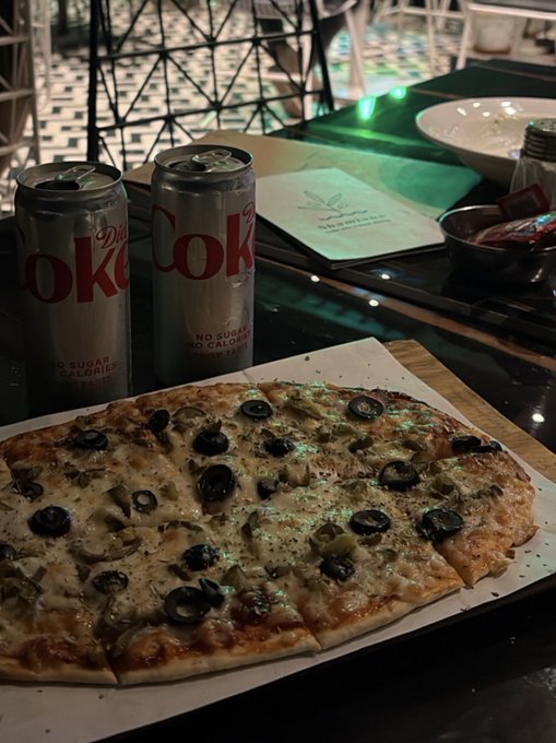 A pizza topped with black olives sits on a wooden board next to two silver Diet Coke cans, with a Hakkasan menu visible on the table.